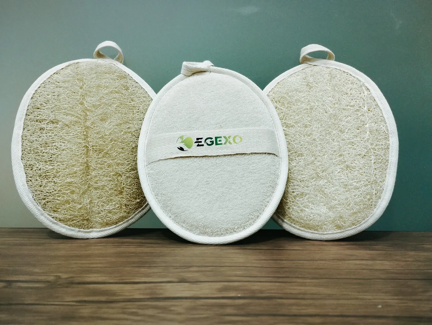 Three oval loofah pads with EGEXO labels standing against a wall on a wooden table