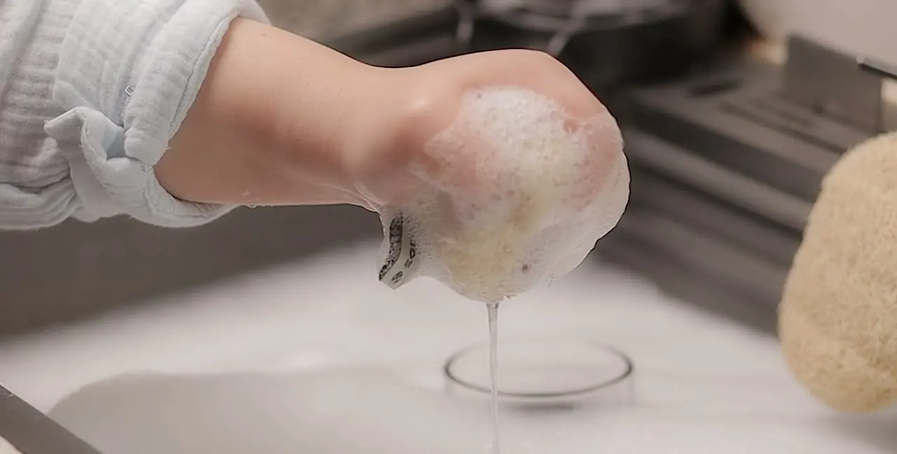 Uma mão espremendo espuma de uma esponja vegetal na pia da cozinha.